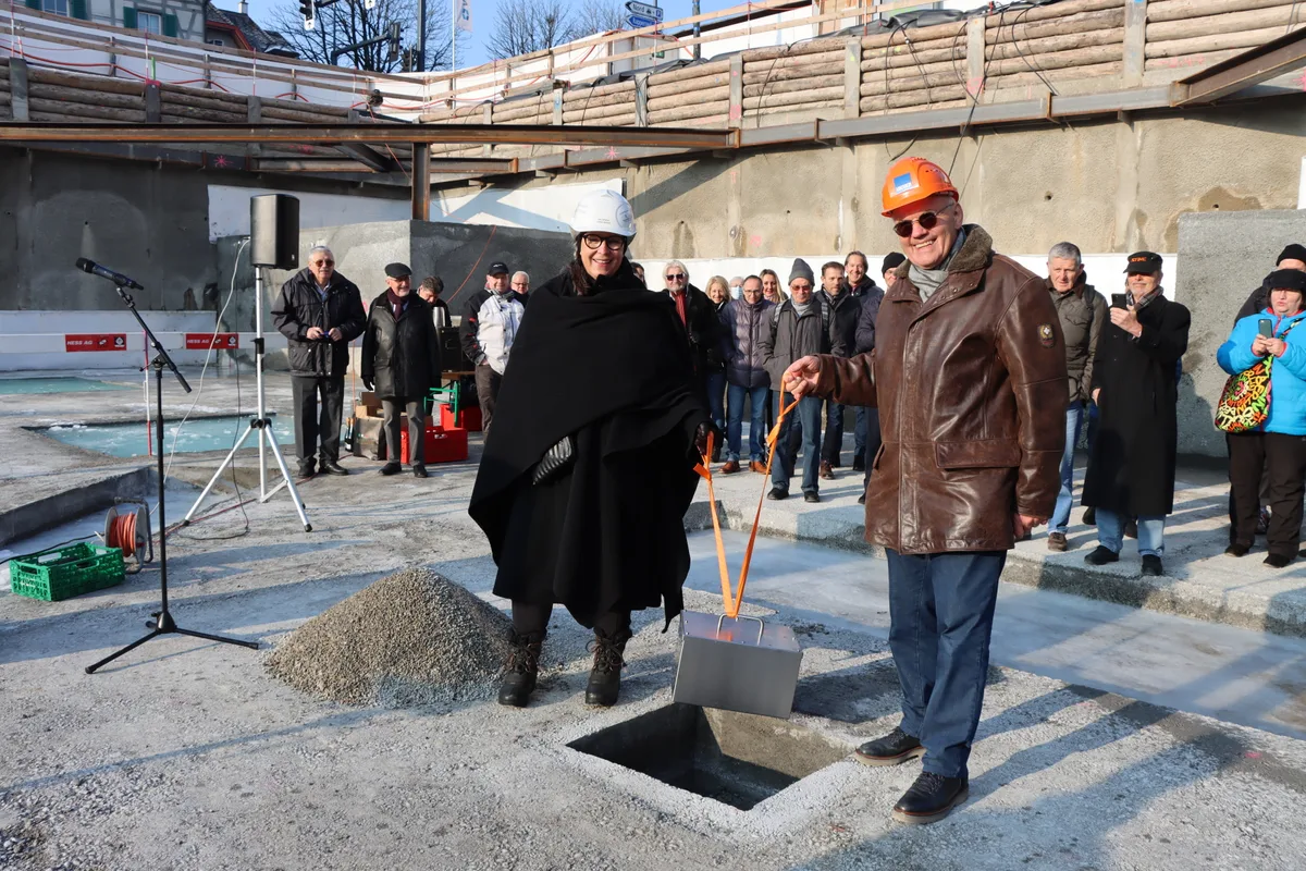 Eine Frau und ein Mann stehen gesichert mit einem Helm auf der Baustelle und halten an einem Band eine Zeitkapselbox über einem Loch im Betonboden.